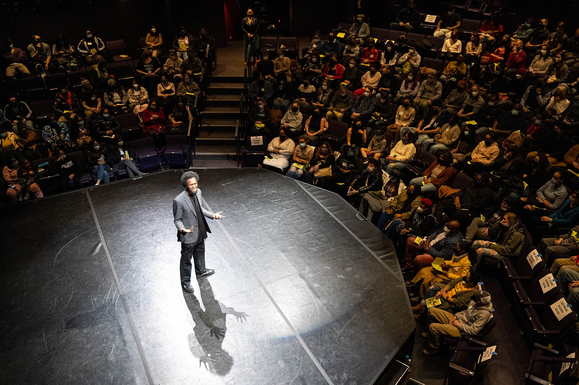 A man stands at the center of a circular stage surrounded by an audience in seats.